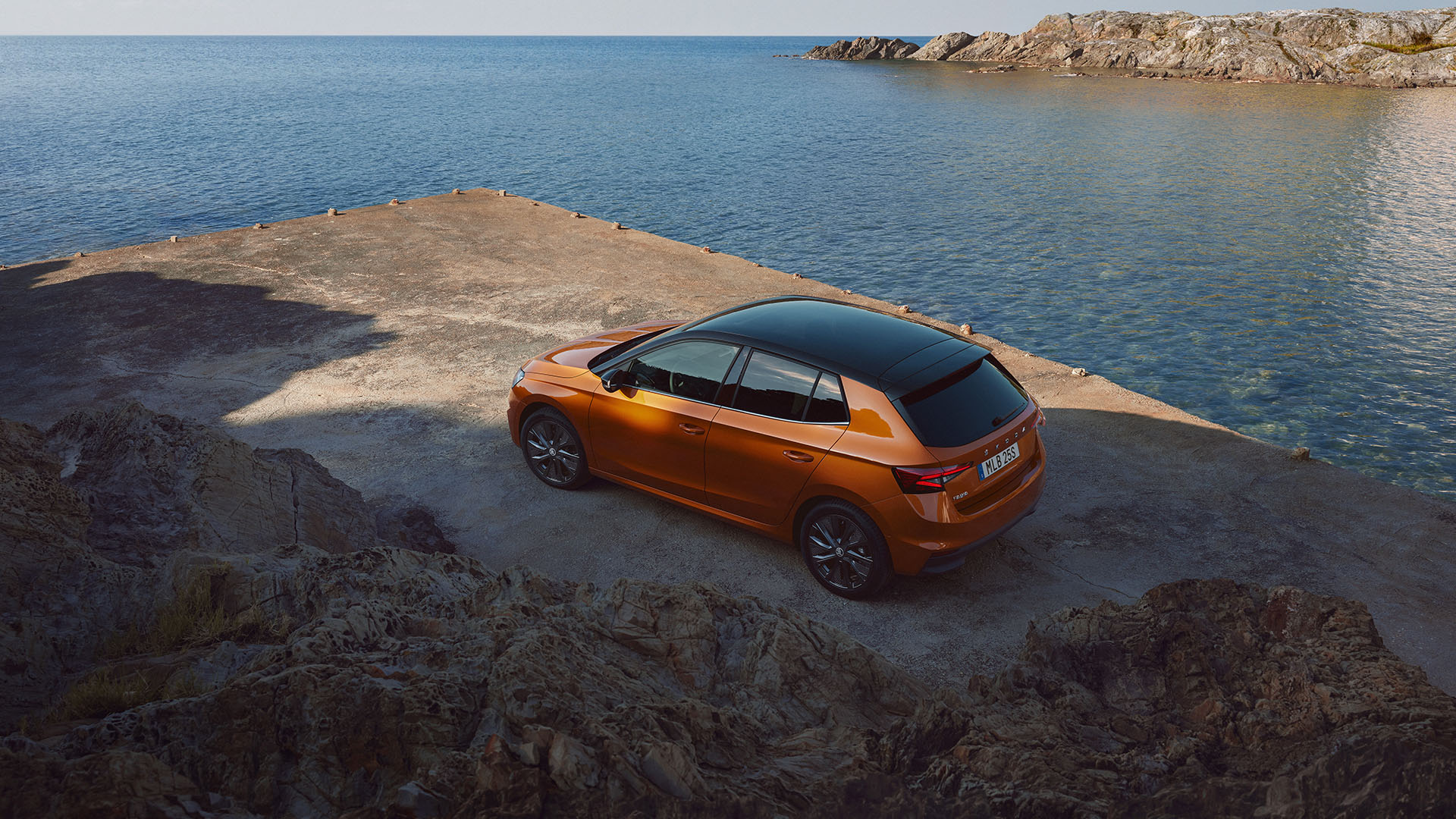 En orange Skoda Fabia med svart tak parkerad ute på en pir med hav och berg i bakgrunden.