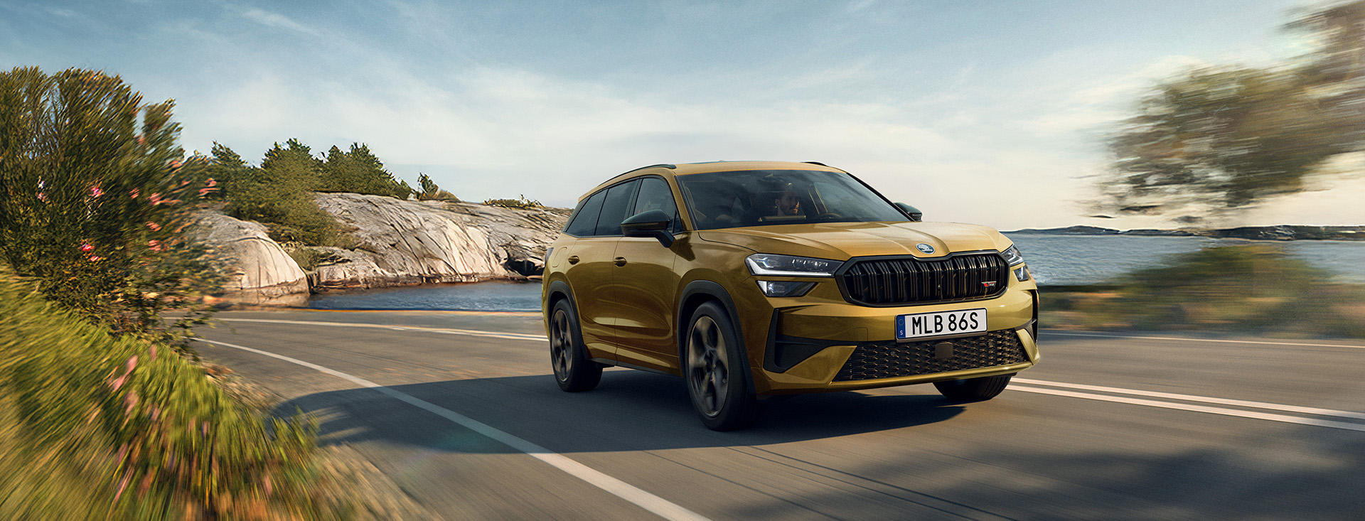 En guldfärgad Skoda Kodiaq kör på väg längs vatten med berg i bakgrunden